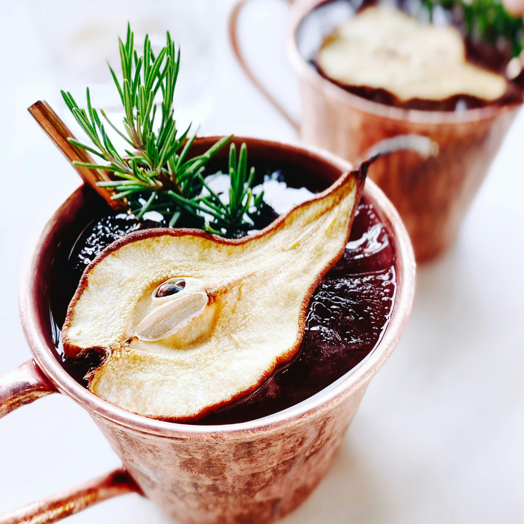 Hot drink in a copper mug with a pear slice and rosemary garnish on a white background