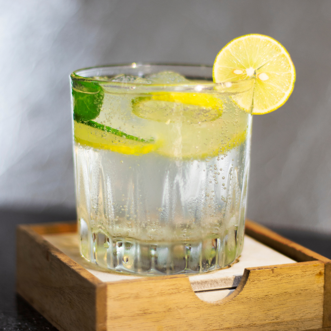 Clear glass of gin, lemonade with lemon slices and a mint leaf on a wooden coaster.